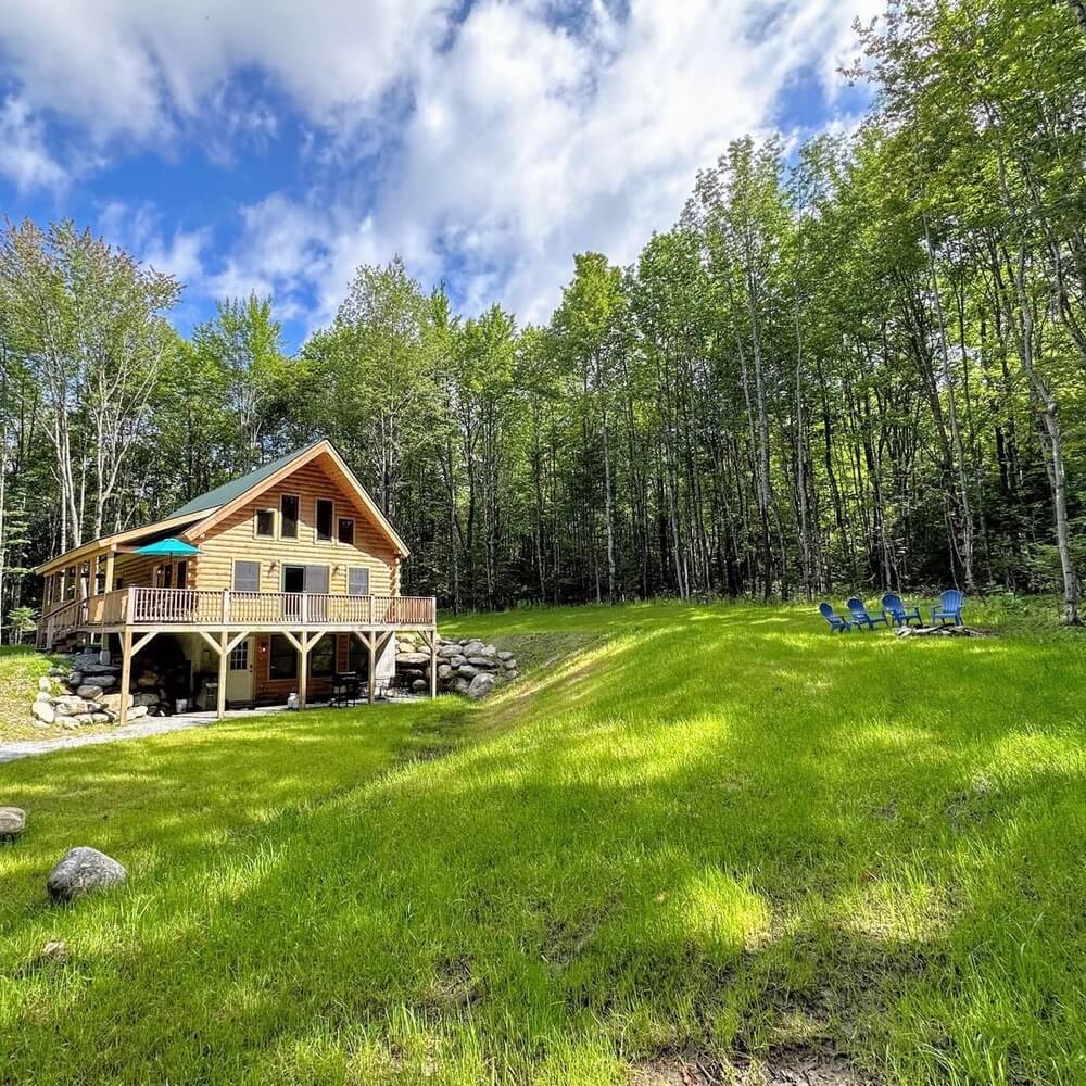 A side view of a large log cabin rental home from Bretton Woods Vacations, featuring a second-story deck with a blue umbrella, a rocky base, and four blue lawn chairs on a grassy hill.